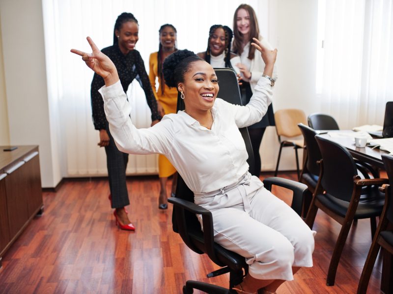 Five multiracial business womans standing at office and roll woman on chair. Diverse group of female employees in formal wear having fun.